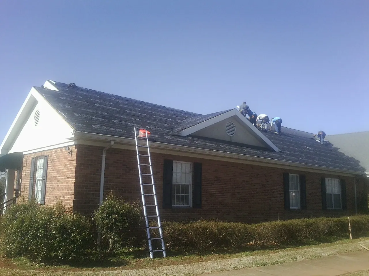 Crew tearing shingles off a brick ranch roof with a ladder set — Emergency Roof Repair work by our crew in Ronan