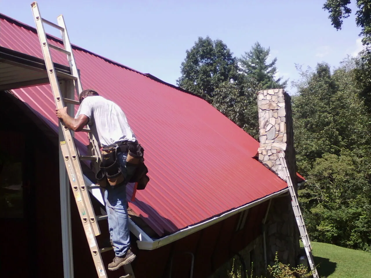 Roofer on a ladder beside a red metal roof and stone chimney — Soffit & Fascia Repair by Roofing Brothers in Ronan